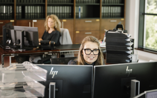 Mitabeiter im Büro, eine Mitarbeiterin schaut freundlich über ihren PC in die Camera